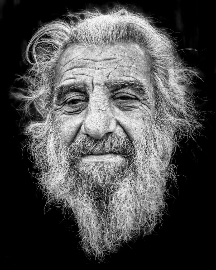Black and white portrait of a senior man with a prominent beard and expressive features against a dark background.